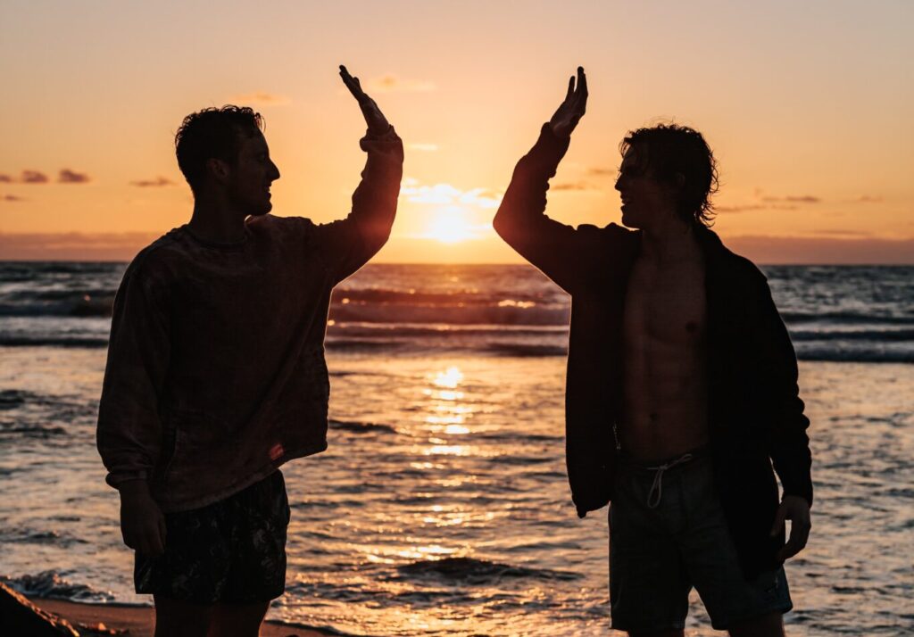 Silhouette of two people high fiving on a beach with a sunrise between them.